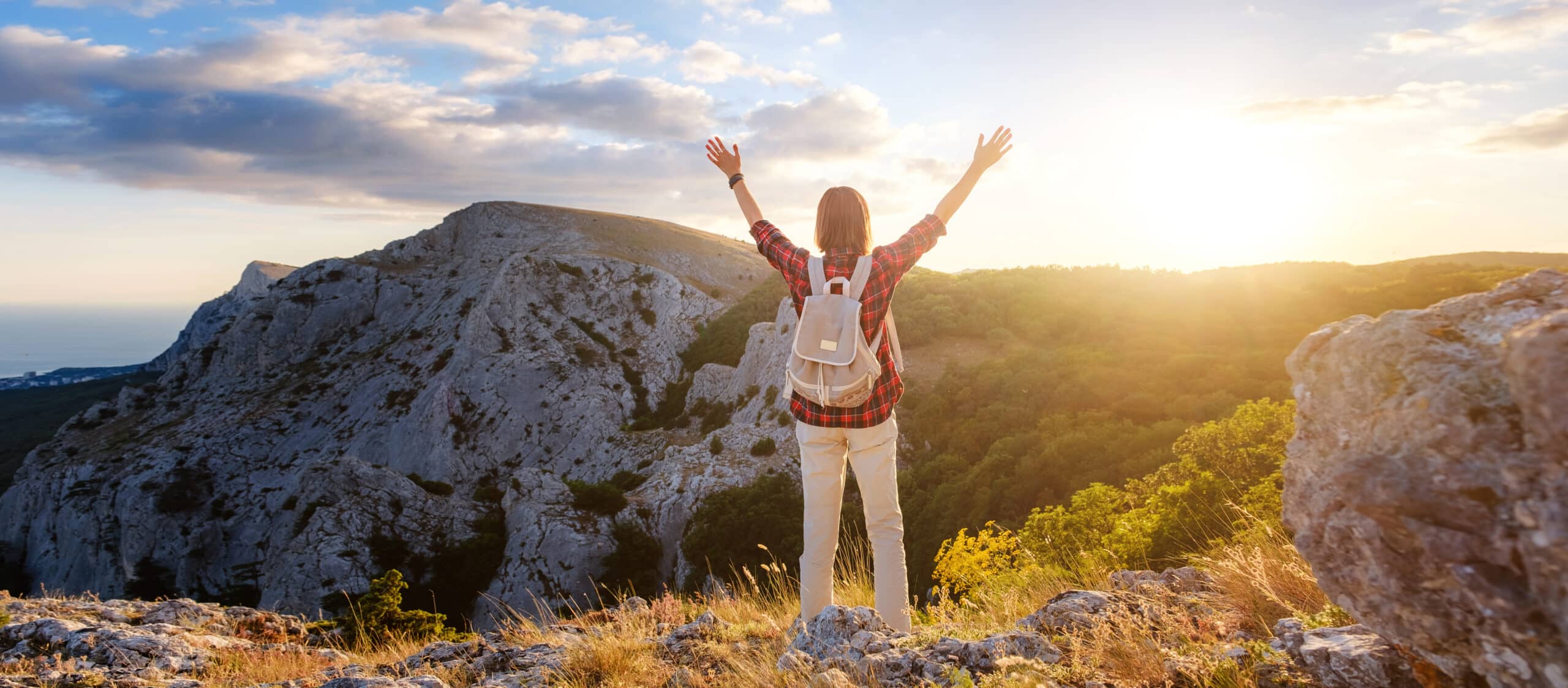 Fit female hiker with backpack standing on a rocky mountain ridge looking out of green alpine valleys and peaks in a healthy outdoors lifestyle concept